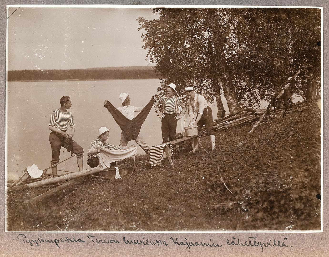 Viisi nuorta miestä pesee vaatteita rannalla kesämaisemassa, taustalla järvi.