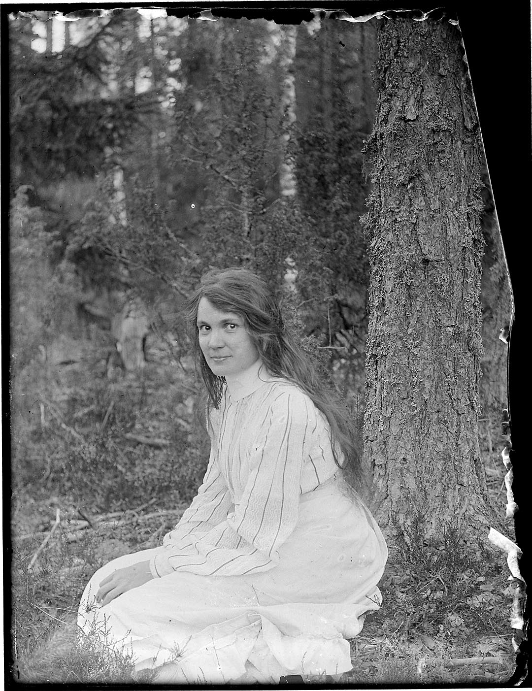 Vaaleaan asuun pukeutunut nainen istuu maassa puun juurella ja katsoo kameraan, pitkät hiukset ovat auki.