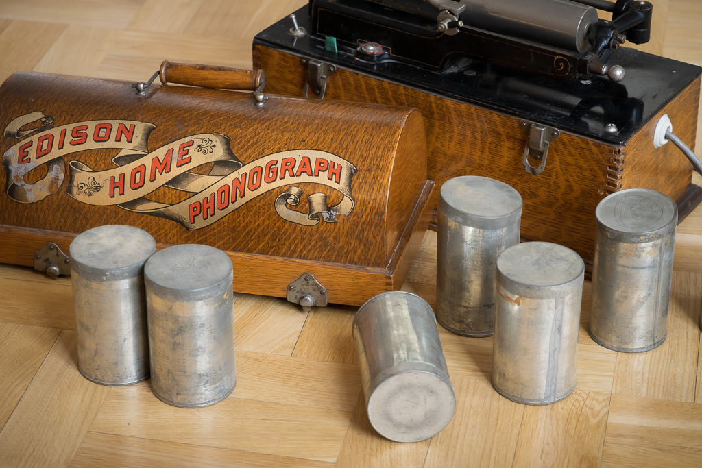Edison Home Phonograph. Trademark Thomas A. Edison. Fonografi parketilla ja vieressä metallisia purkkeja vahalieriöiden säilyttämiseen.