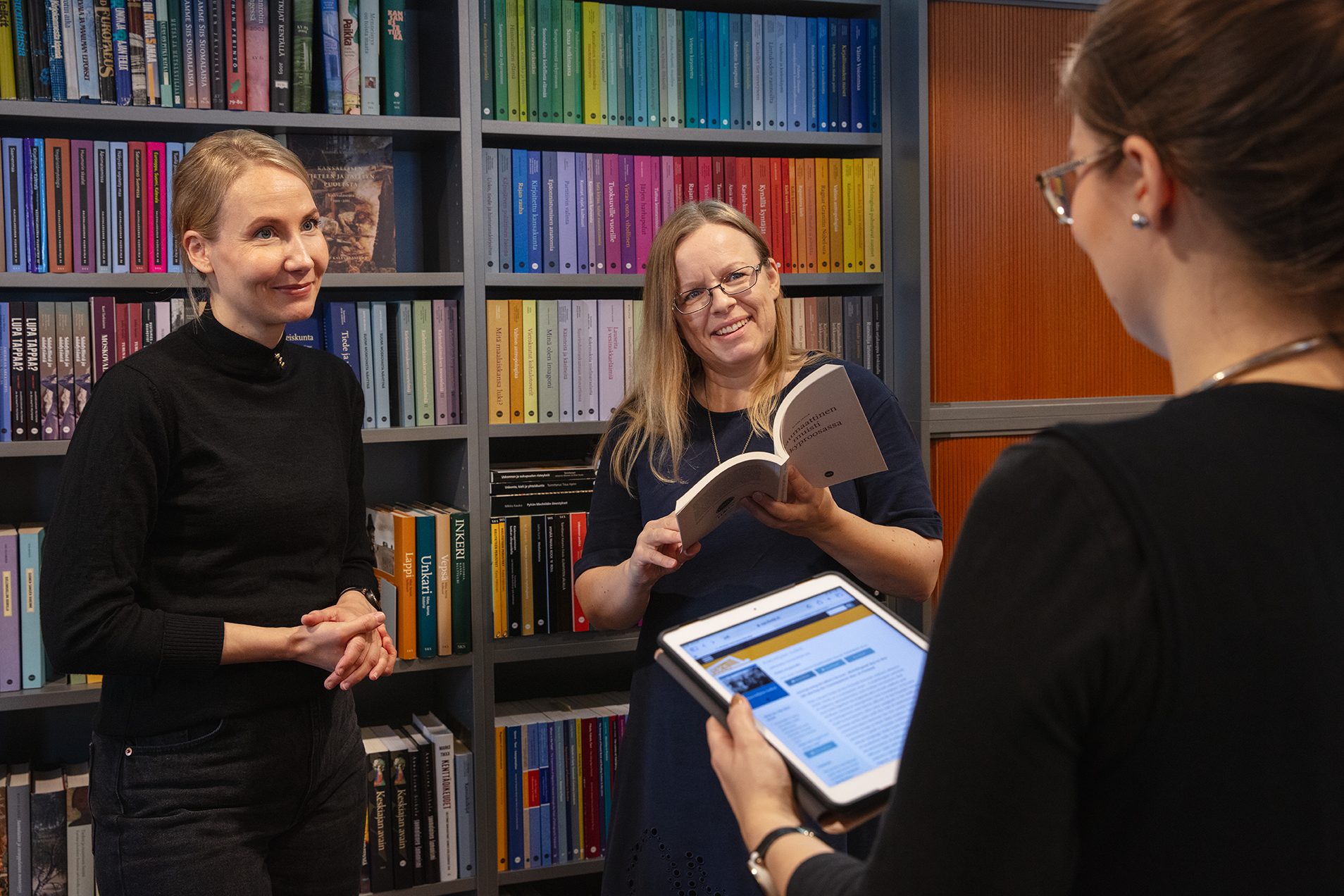 Kolme naista keskustelee kirjahyllyn edessä. Yhdellä on kädessään tabletti, jossa on auki SKS:n avoimien tiedekirjojen julkaisualusta.