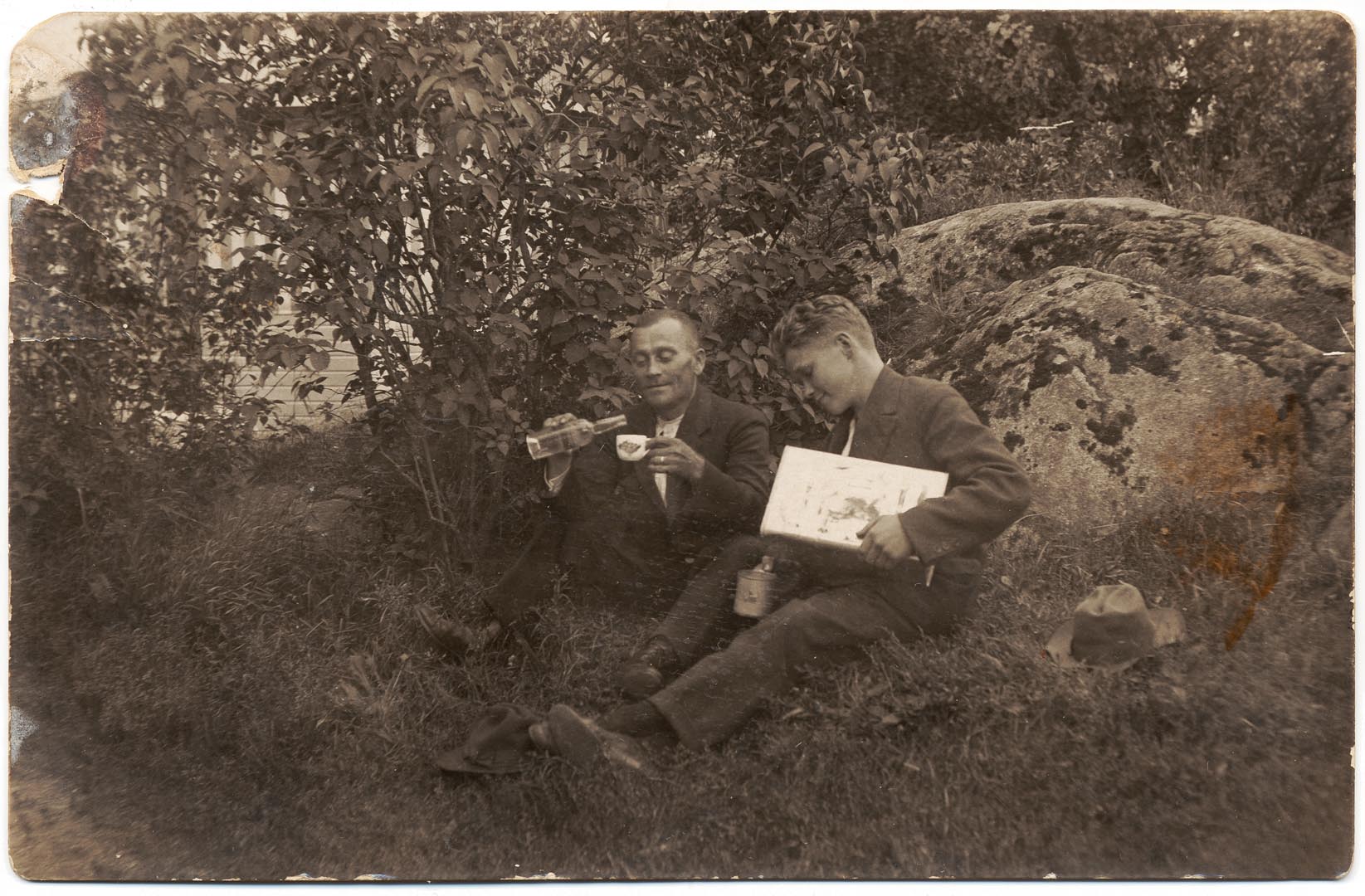 Kaksi miestä istuu kiven juurella juomassa.